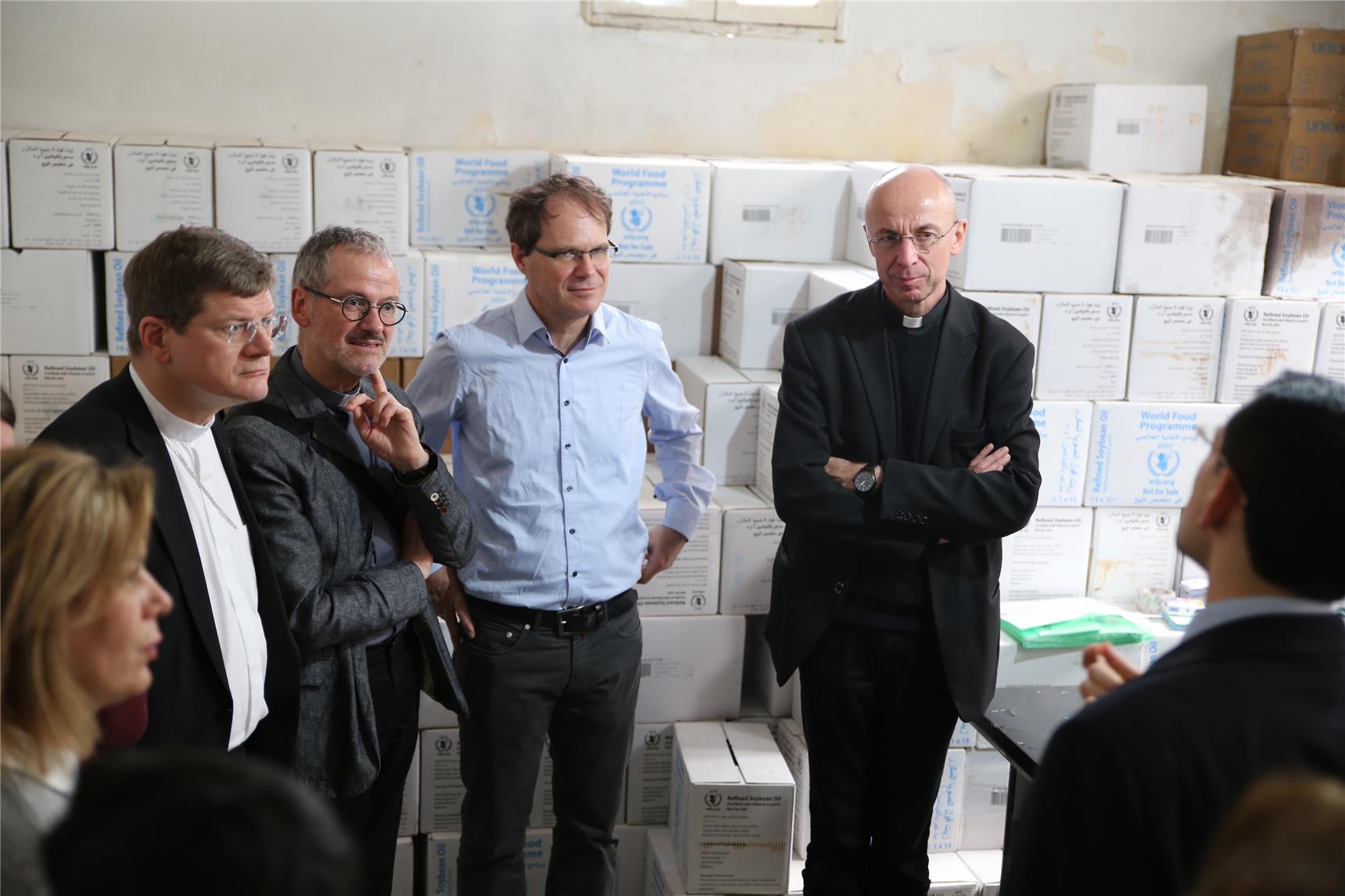 Vier Männer stehen vor einer Zimmerwand, die mit Kisten vollgestellt ist und schauen einen Mann an.