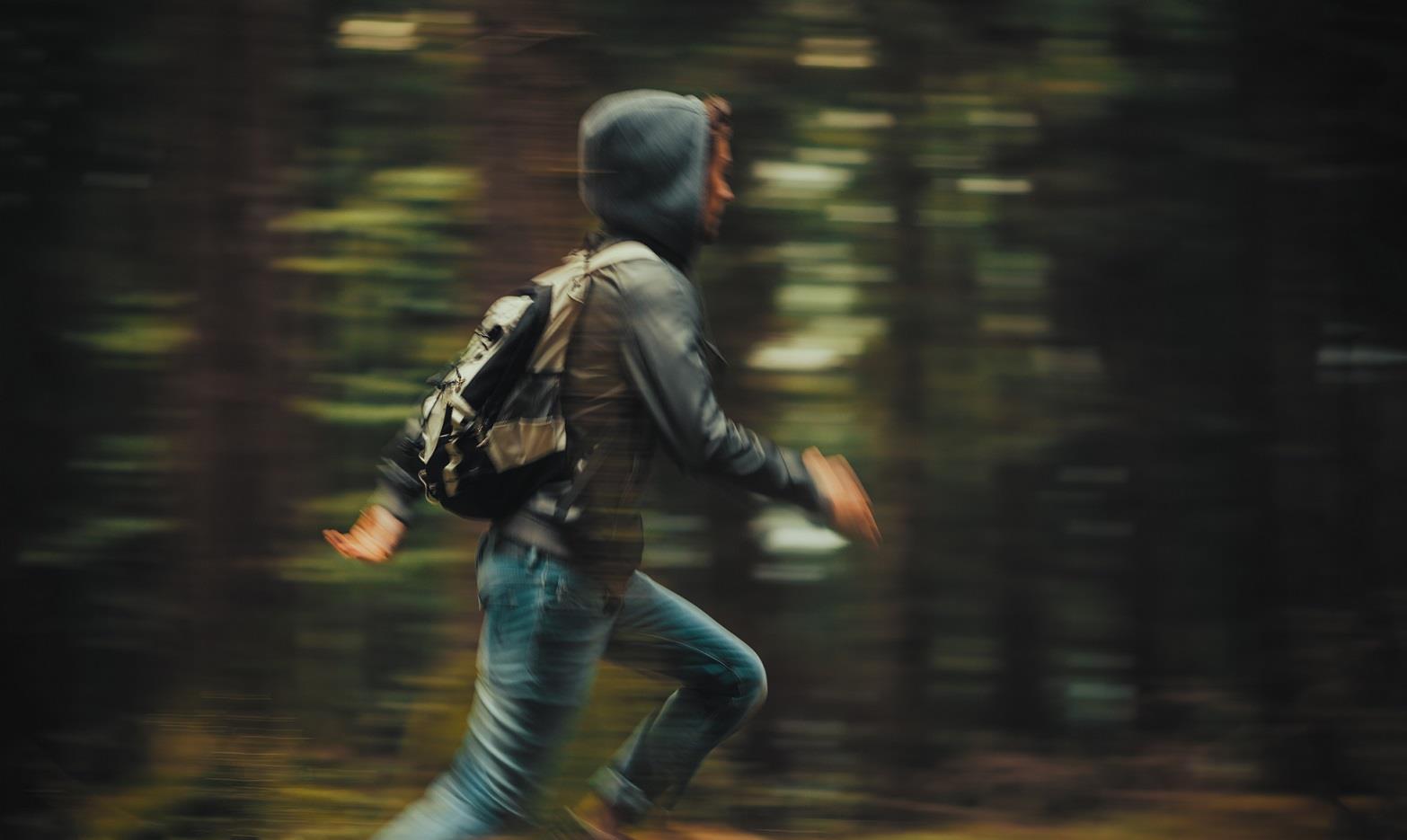 Ein Jugendlicher mit Kapuzenpullover, Rucksack und Jeans rennt durch einen Wald.