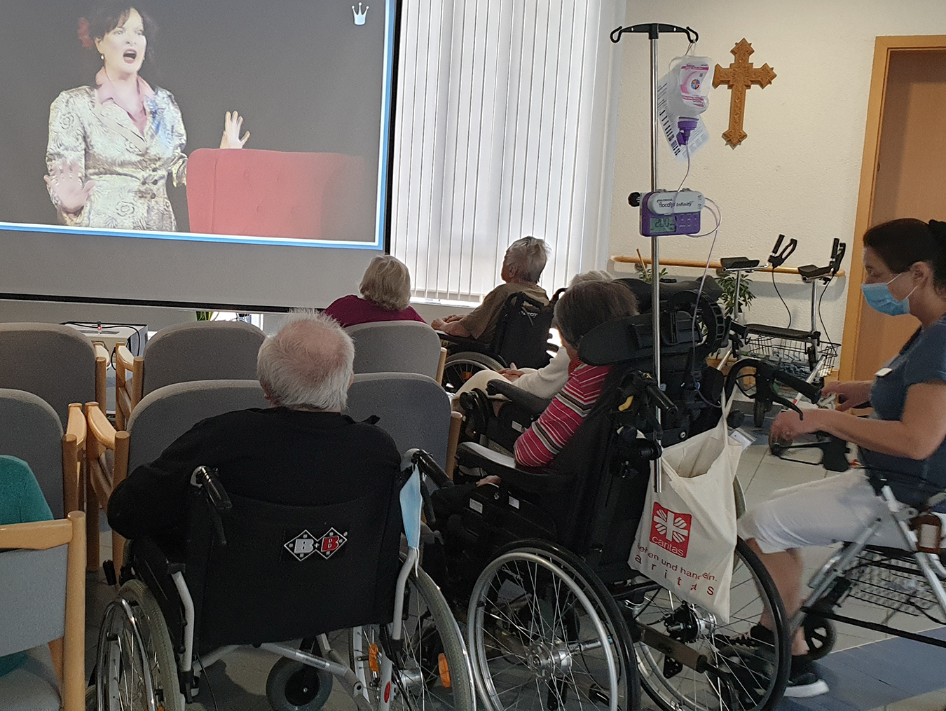 Eine Gruppe älterer Menschen schaut sitzend auf eine Leinwand, auf der eine Frau mit schwarzen langen Haaren und goldenem Kleid zu sehen ist.