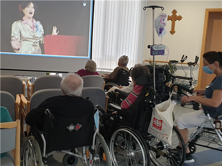Digitale Märchenstunde_Caritas_Bayern Eine Gruppe älterer Menschen schaut sitzend auf eine Leinwand, auf der eine Frau mit schwarzen langen Haaren und goldenem Kleid zu sehen ist.