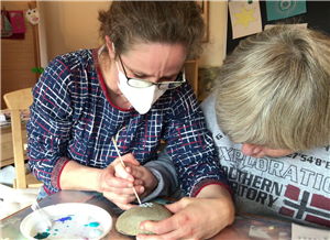 Ausbildung Heilerziehungspflege Heilerziehungspflegerin unterstützt Bewohnerin beim bemalen eines Steins.