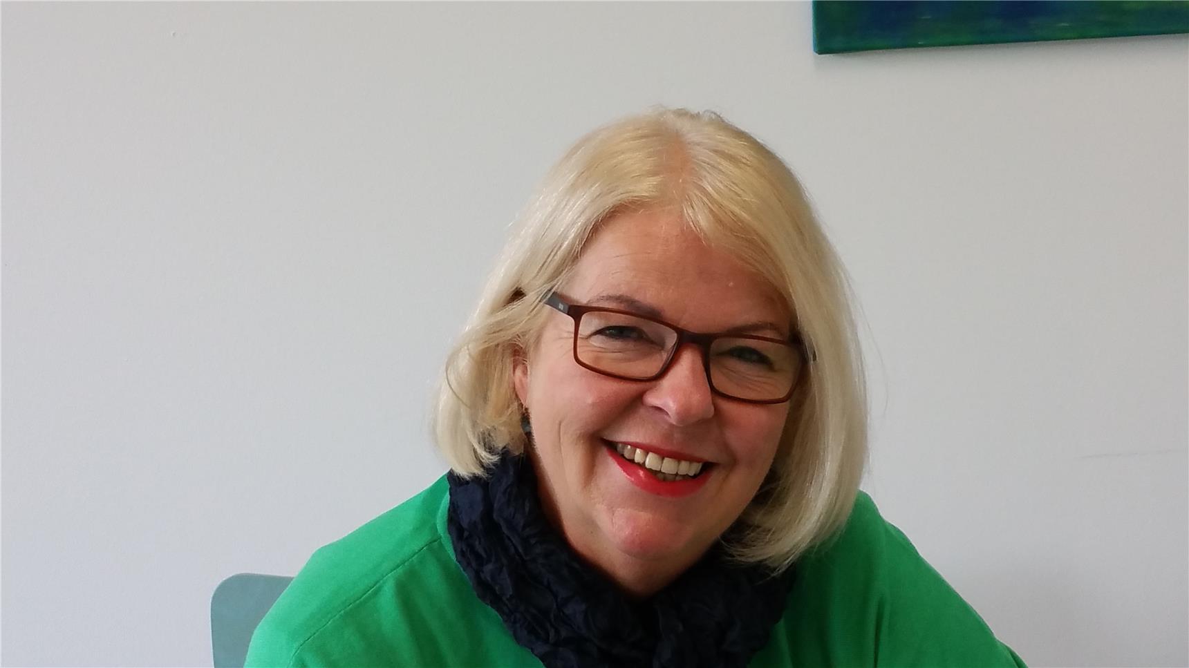 Eine ältere Frau mit mittellangen blonden Haaren und Brille lacht in die Kamera.