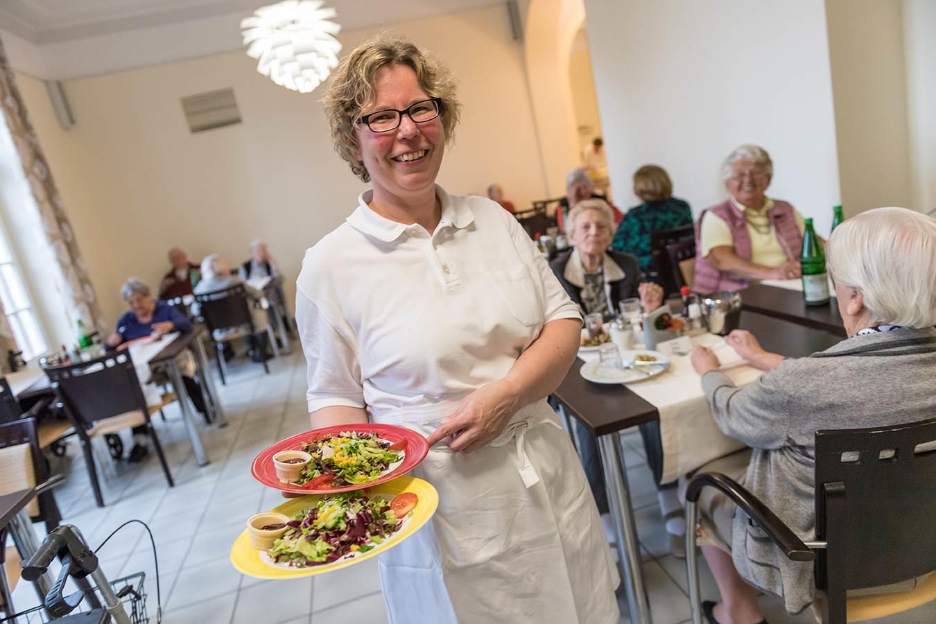 Eine sympathische Hauswirtschafterin bringt Essen in den Speiseraum eines Altenheims. Im Hintergrund Bewohnerinnen an ihren Tischen.