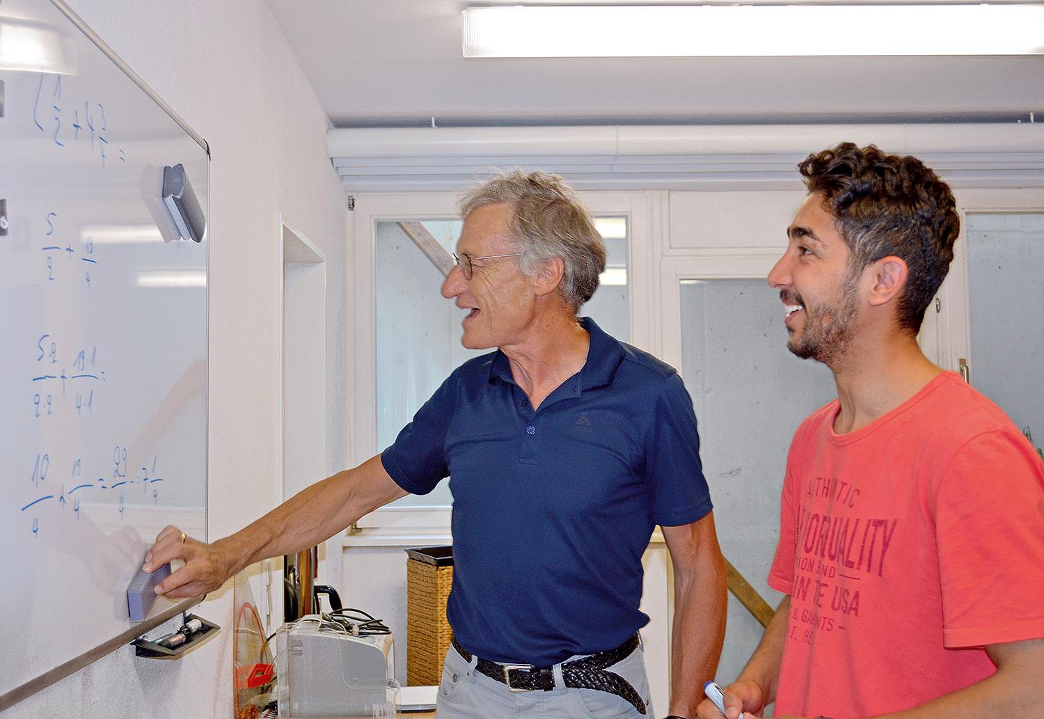 Ein älterer steht mit einem jüngeren Man vor einer Tafel mit Rechenaufgaben, beide lachen.