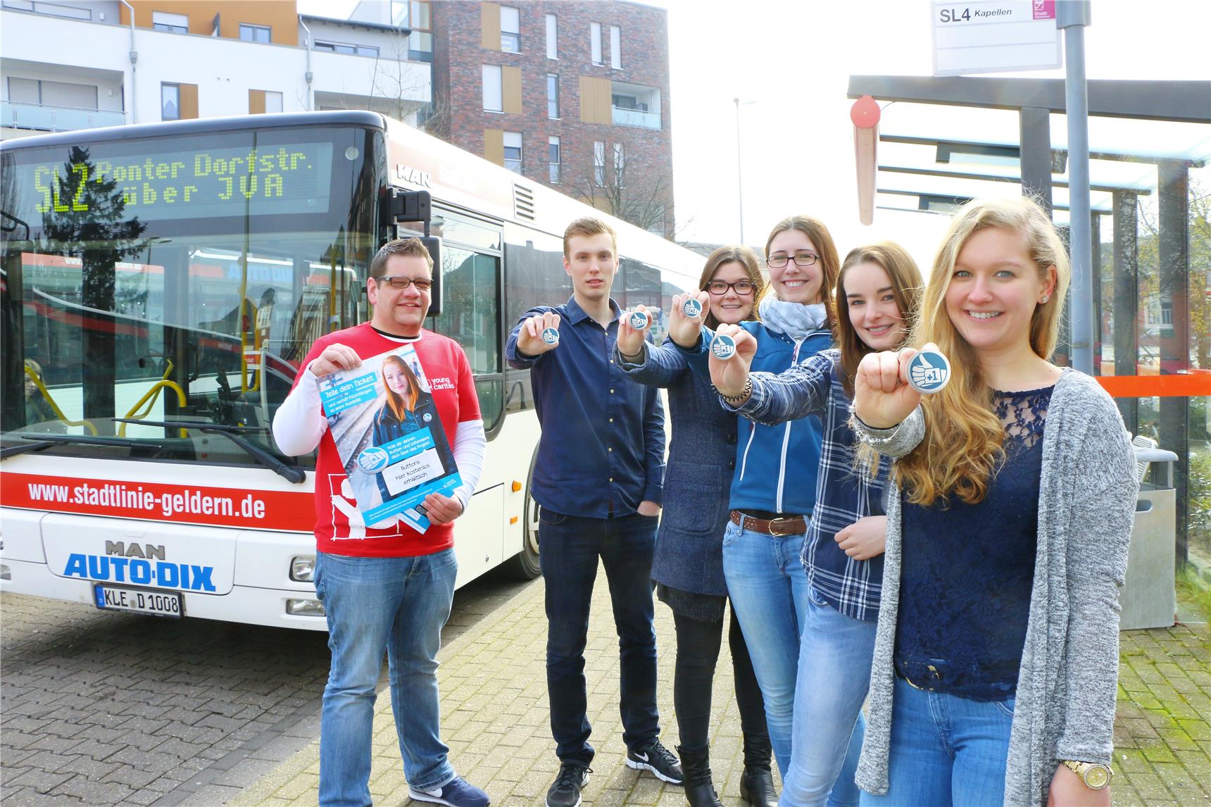 Vier junge Damen und zwei junge Herren stehen vor einem Bus und zeigen blaue Buttons in die Kamera.