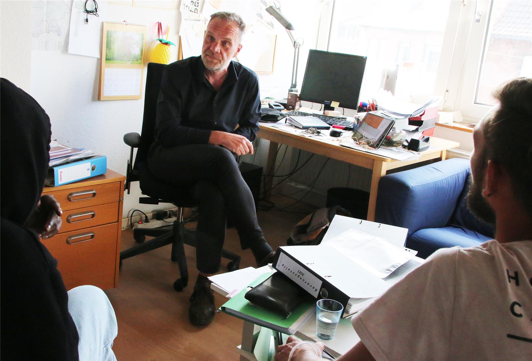 Das Foto zeigt einen Mann vor einem Schreibtisch sitzend im Gespräch mit zwei jungen Erwachsenen.