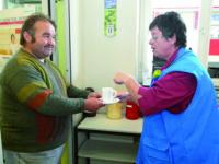 Wir sehen eine ehrenamtliche Helferin, die sich bei einem Kaffee, mit einem Mann in der Bahnhofsmission unterhält. 