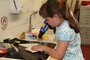 Erfinderwerkstatt_2012_2 Wir sehen ein junges Mädchen mit einem Stück Holz in der Hand. In der anderen Hand hält sie einen Bohrer. Sie scheint viel Spaß bei den Bastelarbeiten zu haben.