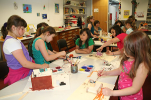 Auf dem Bild sind viele Mädchen zu erkennen, welche ihr Werkstücke mit bunten Farben verzieren. Ein Auto, ein Haus und andere Bastleinen stehen auf dem Tisch und werden bemalt. Die Mädchen scheinen Spaß zu haben.