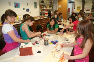 Erfinderwerkstatt_2012_5 Auf dem Bild sind viele Mädchen zu erkennen, welche ihr Werkstücke mit bunten Farben verzieren. Ein Auto, ein Haus und andere Bastleinen stehen auf dem Tisch und werden bemalt. Die Mädchen scheinen Spaß zu haben.