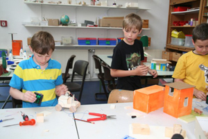 Das Bild zeigt drei Jungen die gerade am Basteln sind. Ein junge hat ein selbstgebautes Holz-Auto vor sich und werkelt daran. Ein anderer Junge schaut ihm dabei zu. Auf der Werkbank stehen noch verschiedenste Sache wie ein Holzhaus und Bastelmaterialien 
