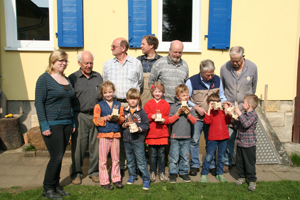 Gemeinsam haben sich die jungen Senioren und die Kinder zu einem Gruppenfoto aufgestellt. Sechs Kinder halten gebastelte helle Holzautos in ihren Händen. Im Hintergrund sehen wir eine Gelbes Gebäude m