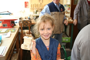 Ida_zeigt_ihr_Auto Ein junges Mädchen hält in ihrer rechten Hand das gebastelte Auto. Auf dem Kühler des Feuerwehrautos ist der Name IDA und ein Logo braun in das Holz eingebrannt. Ida freut sich sehr. Im Hintergrund si