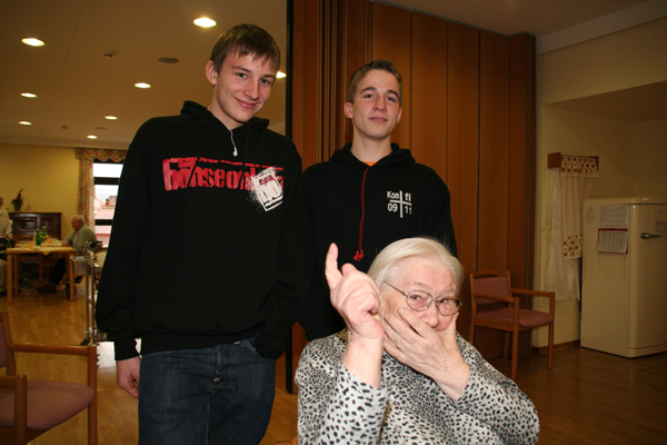 Auf diesem Bild sind zwei Jugendliche mit einer Heimbewohnerin zusehen. Die Bewohnerin hat ihren Zeigefinger in die Luft gestreckt und hat die andere Hand hat sie vor ihrem Mund. Sie scheinen viel Spaß zu haben.Auf diesem Bild sind zwei Jugendliche mit einer Heimbewohnerin zusehen. Die Bewohnerin hat ihren Zeigefinger in die Luft gestreckt und hat die andere Hand hat sie vor ihrem Mund. Sie scheinen viel Spaß zu haben.