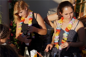 WG_Huenfeld Wir sind zu Gast bei einem Fest. Zwei junge Frauen haben stehen an einem sonnigen Tag hinter einer Bar. Sie mixen Getränke für Gäste, die von ihnen bedient werden. Sie sind in ihre Arbeit vertieft und sprechen mit ihren Kunden.