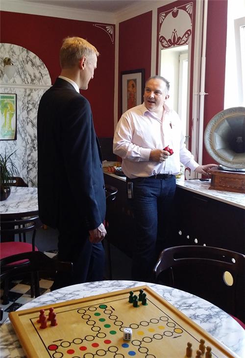 Wir treffen 2 Männer in einem bewusst altertümlich eingerichtetem Zimmer. der eine Mann steht uns zugewandt am Grammophon und spricht mit seinem Gegenüber. Schwarz-weiße Marmorplatten wurden als Ablag