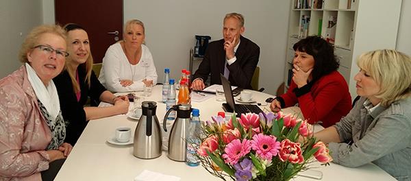 Wir sind zu Gast bei dem Audit im Altenpflegezentrum in Kassel. 6 Personen sitzen um einen großen weißen Tisch. Vor ihnen liegen Arbeitsmaterialien und ein Laptop. Am Ende des Tisches ist ein großer b