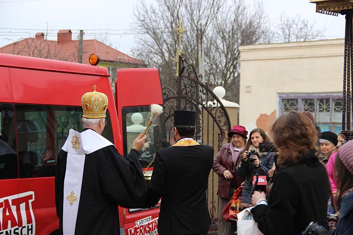 Der Ortsbischof im schwarzen Talar und weißer Stola schwenkt den im Weihwasser getauchten Aspergill. Er segnet damit den roten Caritasbus aus Deutschland. Der Fortbus trägt die Aufschrift: Caritas Werkstatt Mensch.