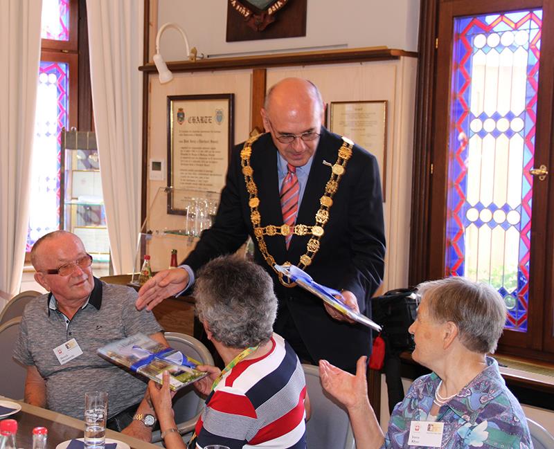 Unsere polnischen Gäste sind im Rathaus zu Besuch. Der Bürgermeister verteilte gerade eine Mappe oder Buch an die Teilnehmer. Dabei wendet er sich direkt an jeden Gast. Er trägt auf seinem Jackett eine goldene Kette. 