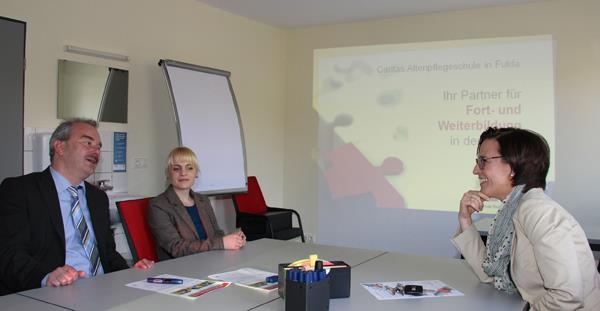 In einem hellen Raum sind mittig Tische zu einer großen Arbeitsfläche zusammengestellt. Auf ihm liegen Büromaterialien wie Stifte und Notizblöcke. Drei Personen sind auf den forderen Plätzen zu sehen.