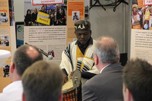 Vor den Ausstellungplakaten spielt ein dunkelhäutiger Afrikaner in seiner Nationaltracht eine selbst gebaute Trommel. Viele Gäste haben sich vor ihm versammelt und schauen ihn an.