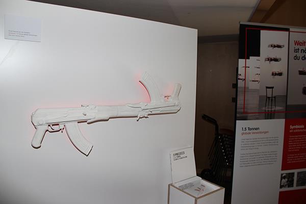 Auf einem weißen Hintergrund sind zwei weiß gestrichene Maschinengewehre waagerecht so angebracht, dass die Läufe gekürzt und entgegengesetzt zusammengeschweißt wurden. Der Schuss des einen Gewehres würde im Lauf des anderen Gewehres enden. 