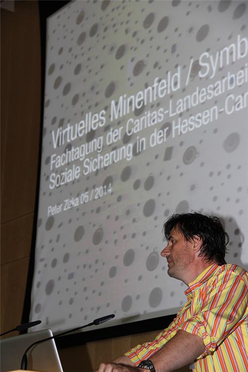 Peter Zizka steht am Rednerpult. Hinter und neben ihm ist eine große Leinwand zu sehen auf grauem Hintergrund sind wiederum graue Kreise zu dargestellt. Das Thema 'Virtuelles Mienenfeld' wird in weiße