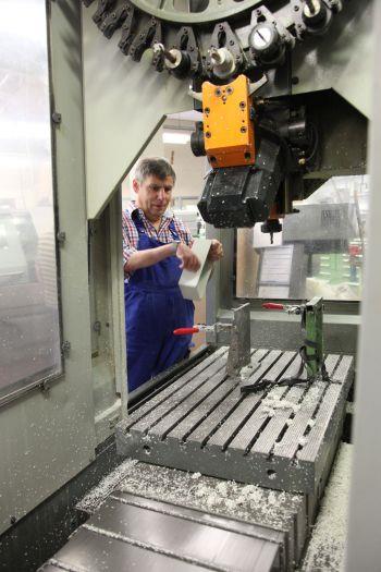 Wir blicken von hinten in eine CNC-Maschine. Ein Mann hat gerade ein weißes Formteil aus Plastik ausgespannt und hält es in seinen Händen. Auf dem Schlitten der Maschine liegen überall weiße Späne