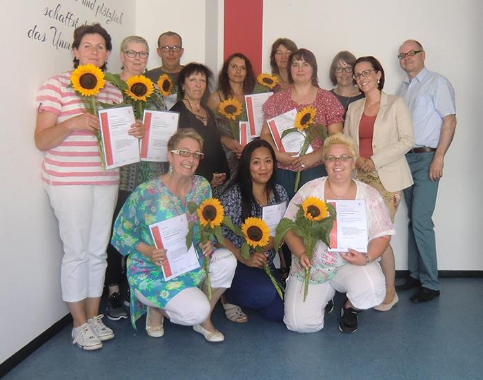 Die Betreuungskräfte und ihrer Ausbilder haben sich als Gruppe in einer Ecke der Altenpflegeschule zusammengestellt. Die Absolventen haben je eine Sonnenblume in ihrer Hand. 