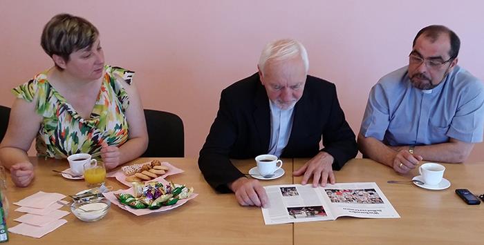Wir sehen den Bischof wie er in der Zeitschrift blättert. Neben ihm sitzen ein Priester und eine Frau.
