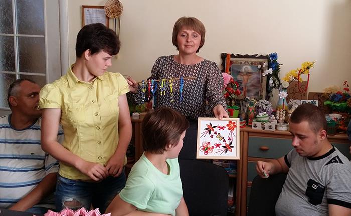 Wir sind bei einer Familie zu Gast. Die Mutter hält in der rechten Hand gestaltete Armbänder und in der linken Hand ein Bild mit Blumen und einem Schmetterling. 