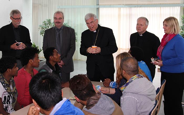 Wir sind zu Gast in der Flüchtlingsunterkunft in Neuhof. Der Weihbischof und Vertreter des Caritasverbandes stehen im Plenum vor vielen Tischen, an denen Flüchtlinge sitzen. 