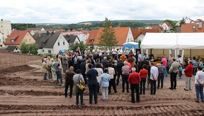 Viele Menschen aus Politik, Kirche, dem Bauhandwerk, des Caritasvorstandes und Betroffene haben sich auf der Baustelle vor einem Zelt um ein Rednerpult versammelt. Im Hintergrund spielt eine Blaskapelle ein Eröffnungslied.
