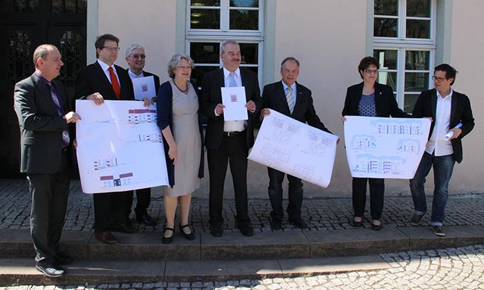 Die Überbringer und Empfänger der Förderbescheide haben sich vor die Geschäftsstelle des Caritasverbandes in Fulda gestellt. Sie halen Zeichnungen von den Wohnprojekten wie Banner vor sich!