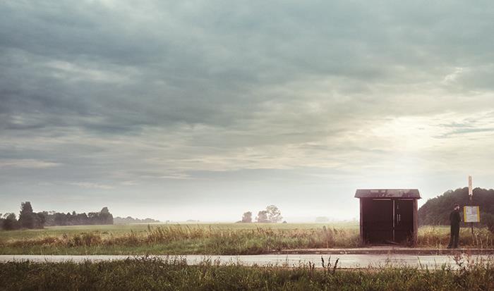 Ein tristes Landschaftsbild zeigt an einer Straße in der rechten Ecke eine vereinsamte kleine Bushaltestelle. Diese ist vor vielen Jahren aus Brettern zusammengebaut. Neben dem Häuschen steht ein Mann