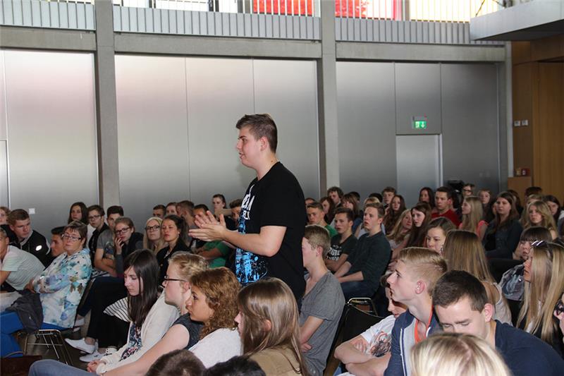 Wir blicken auf die zuhörenden Schüler des Marianums. Ein Schüler steht in der Menge und hält seine Arme nach vorn. Er stellt sicherlich eine Frage.