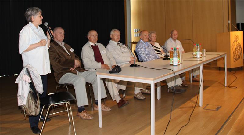 Die sechs Zeitzeugen sitzen im Plenum an zwei langen Tischen. Eine Dolmetscherin steht links neben den Tischen und spricht in ein Mikrofon. 