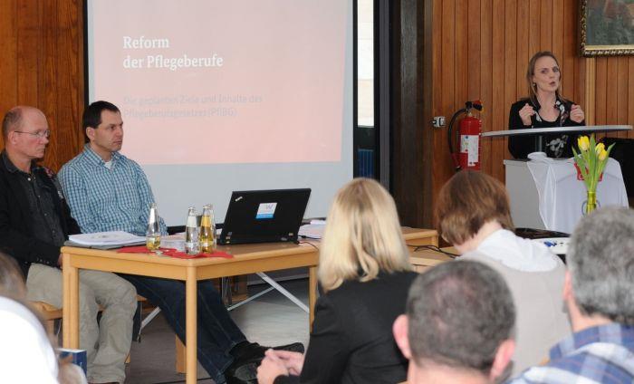 Wir blicken über die Teilnehmer der Infoveranstaltung in das Präsidium. Frau Giesel steht hinterm Rednerpult. Auf einer Leinwand wird über das Thema informiert. 