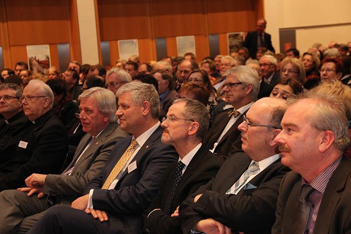 Wir blicken auf die erste Reihe der Teilnehmer. Gespannt schauen sie nach vorn. Im Hintergrund sehen wir weitere Gäste. 