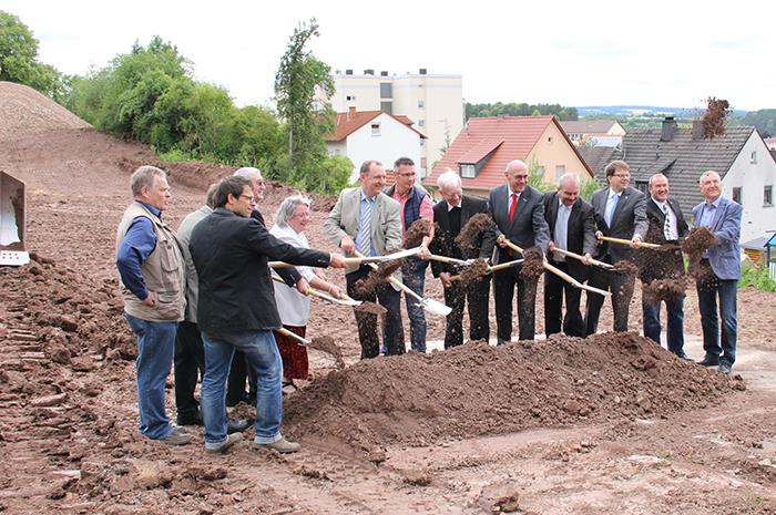 Die Verantwortlichen aus Politik, dem Bauhandwerk, dem Bistum und der Caritas haben sich in einer Reihe hinter einem Erdhügel aufgestellt. Sie haben je einen Spaten und werfen gleichzeitig eine Schauf