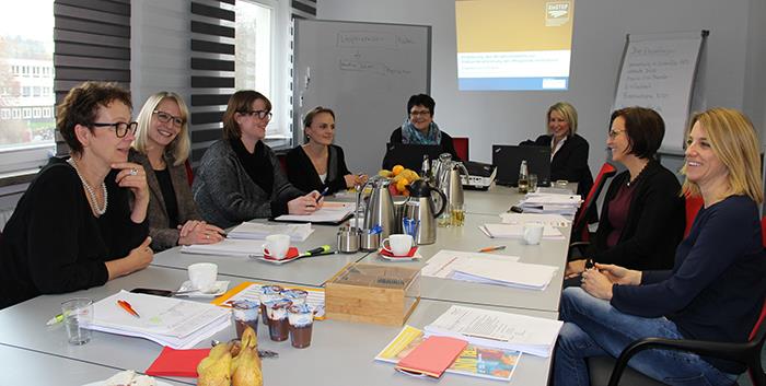 Wir sind zu Gast in der Schulung. An einem großen weißen Tisch haben viele Frauen Platz genommen. Im Hintergrund wirft ein Beamer eine Bild an die Wand. 