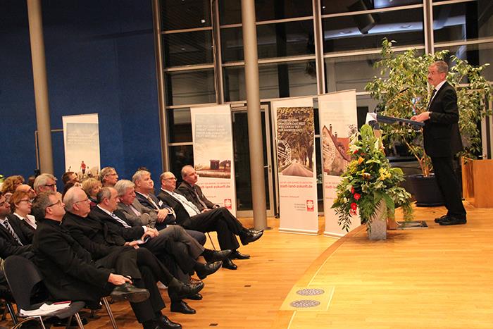 Wir blicken schräg in den Versammlungsraum. Rechts steht Herr Dr. Neher am Rednerpult. Interessiert schauen die Anwesenden zu ihm.