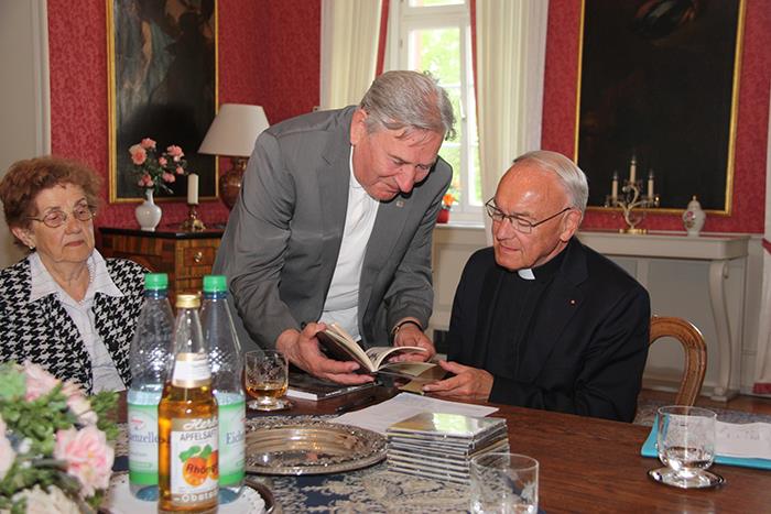 Ein Mann steht neben dem sitzenden Bischof, welcher in einem Buch blättert.