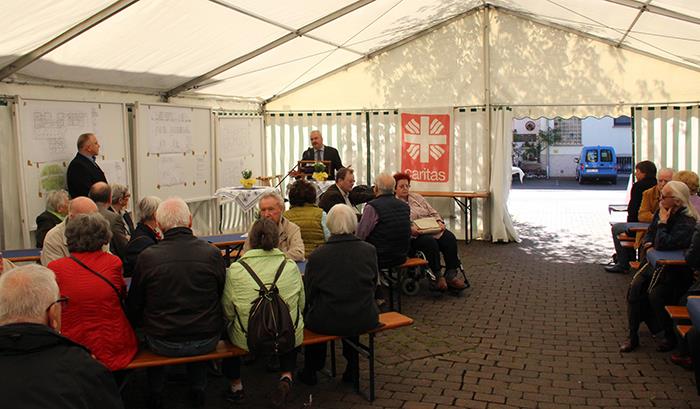 Im Festzelt – Ansprache Caritasdirektor Juch. Wir blicken zum Eingang eines Festzeltes. Hier ist ein Rednerpult aufgebaut. Unser Caritasdirektor steht am Mikro. Links neben ihm ist ein Tafel mit verschiedenen Plänen aufgehangen.