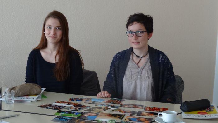 Zwei junge Frauen sitzen hinter einem Schreibtisch und schauen uns an. 