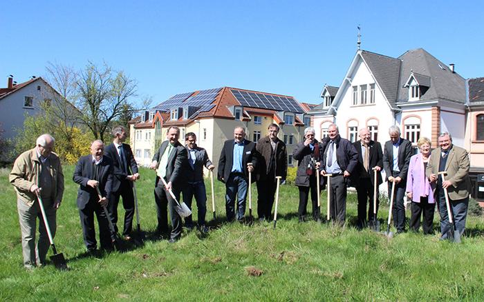 Die Verantwortlichen für das Projekt stehen im Halbkreis auf einer grünen Wiese. Sie halten je einen Spaten in der Hand und stechen die ersten Grasnarben ab.