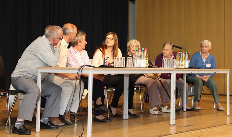 Auf der Bühne der Aula im Marianum sitzen hinter 2 Tischen die 6 Zeitzeugen. Die 3 Herren auf der linken Seite blicken zu einer jungen Frau in der Mitte. Sie schaut zu den Herren. Wahrscheinlich ist es die Dolmetscherin, die gerade etwas übersetzen will. Auf der rechten Seite sitzen 3 polnische Damen, welche auch zu den Männern schauen. 