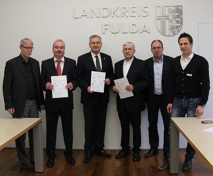 Die Vertreter des Landkreises und der Caritas stehen vor einer weißen Wand mit dem Logo des Landkreises. Die Herren Juch, Woide und Gurk halten ein Papier in ihren Händen. 