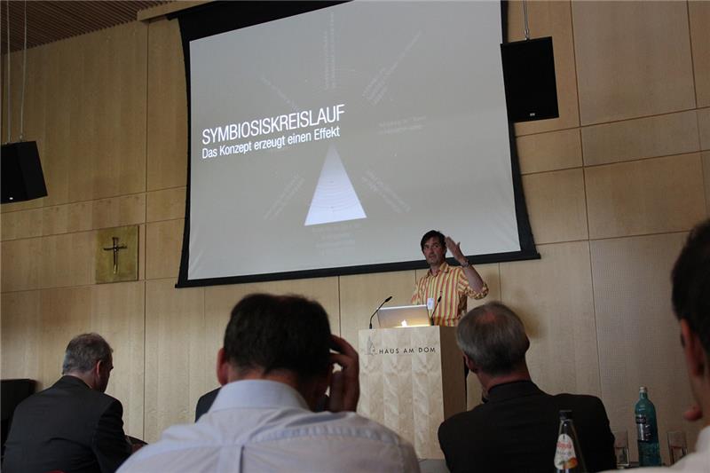 Wir blicken aus der Sicht der Teilnehmer zum Podium. Peter Zizka referiert rechts am Retnerpult. Er hebt seinen linken Arm. Über ihm ist eine Folie seiner Präsentaton zu sehen. Symbiosiskreislauf - Das Konzept erzeugt einen Effekt ist in Heller Schrift über einem hellen Dreieck auf grauem Hintergrund zu lesen. 