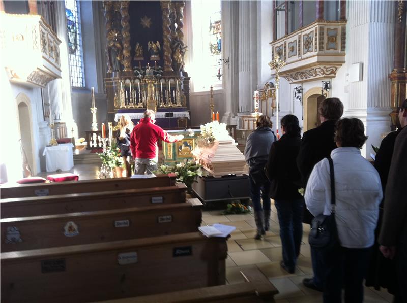 Menschen geben einem Toten die letzte Ehre. Sie treten vor einen Sarg und segen den Toten mit Weihwasser.